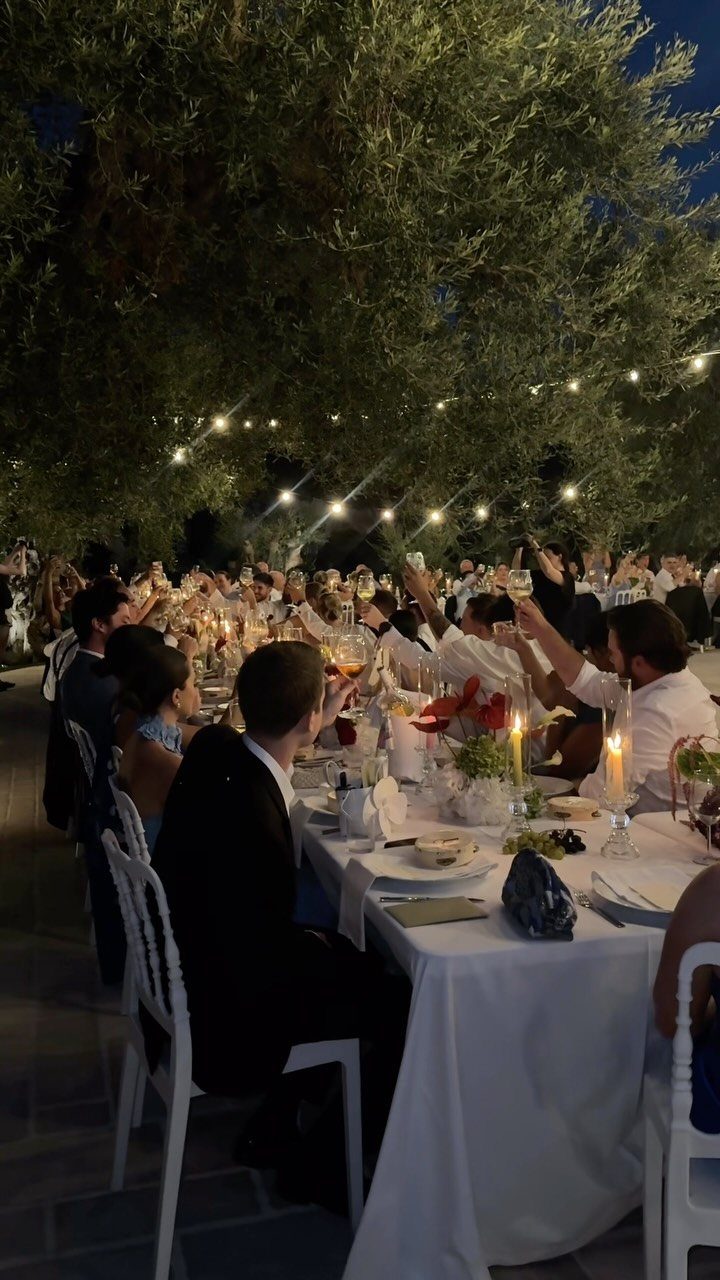 Wedding guests raising glasses for a toast at long white tables under string lights.