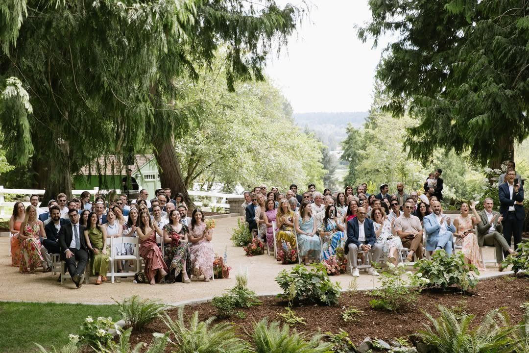 Wedding guests seated in white chairs outdoors under lush green trees.