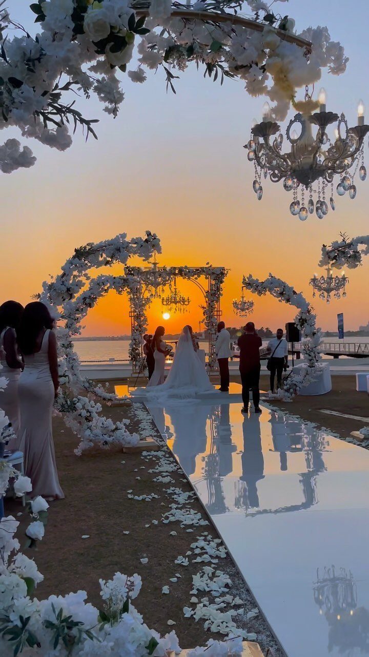 Wedding ceremony at sunset with white floral arches and reflective white aisle.