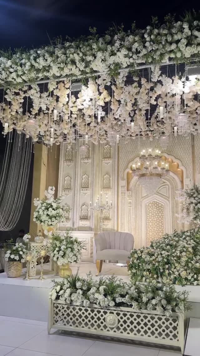 Ornate wedding stage with white flowers, a grey armchair, and carved cream architecture.