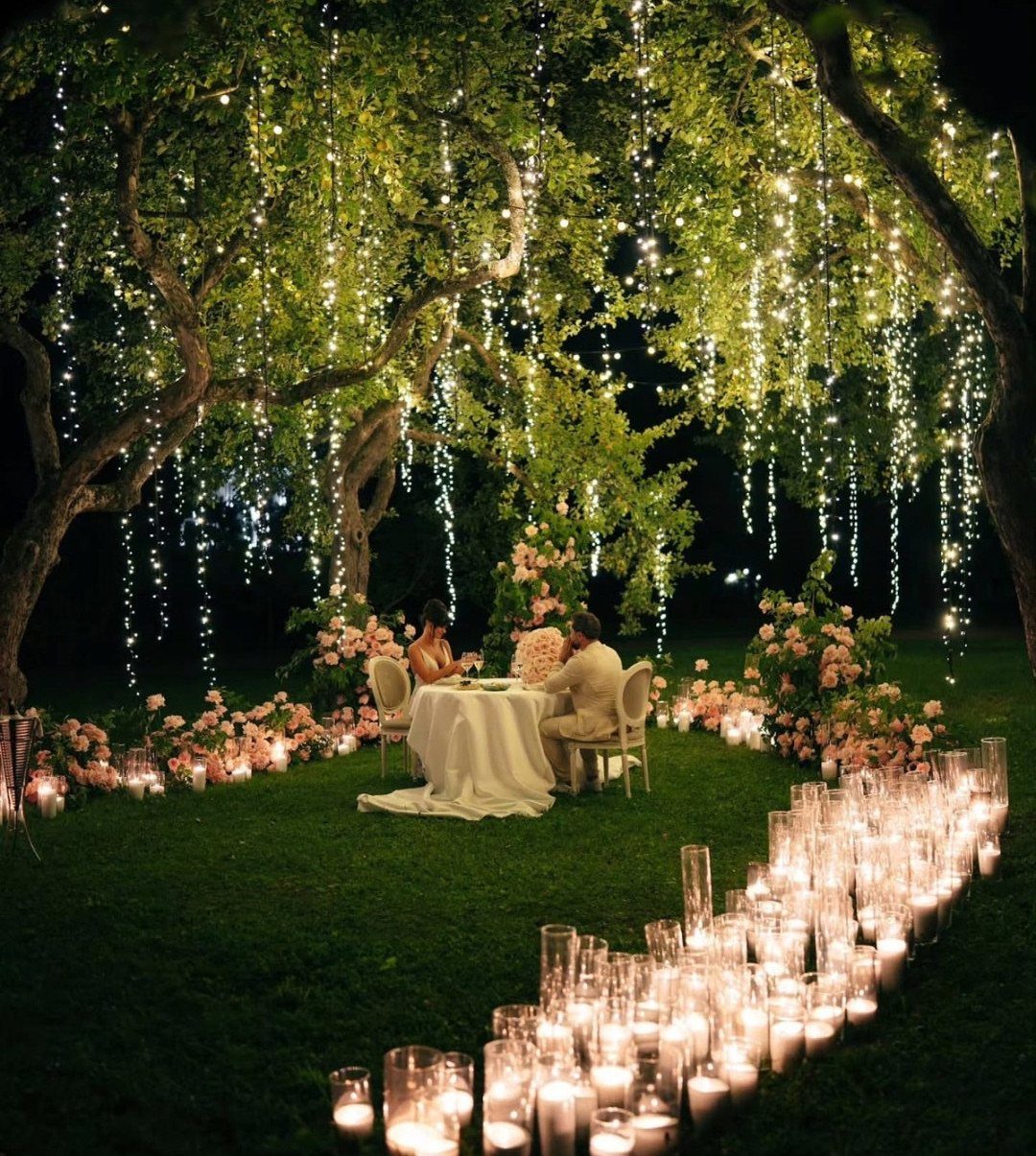 Couple dining under a tree with hanging fairy lights and candlelit grass path