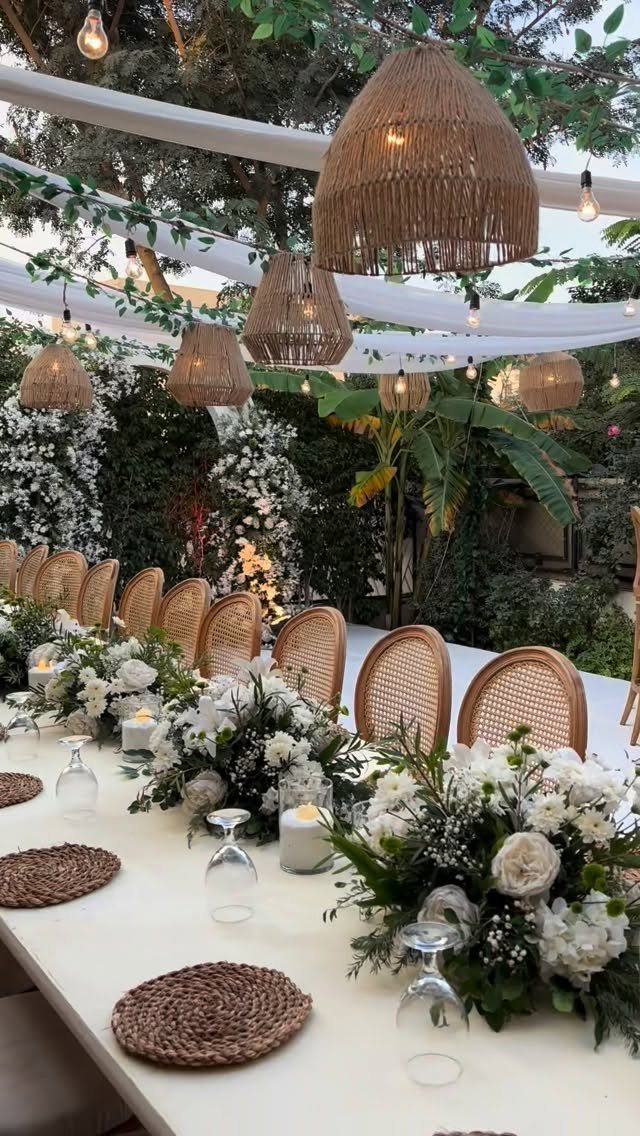 Outdoor wedding table with white flowers, woven chairs, and hanging straw lamps.