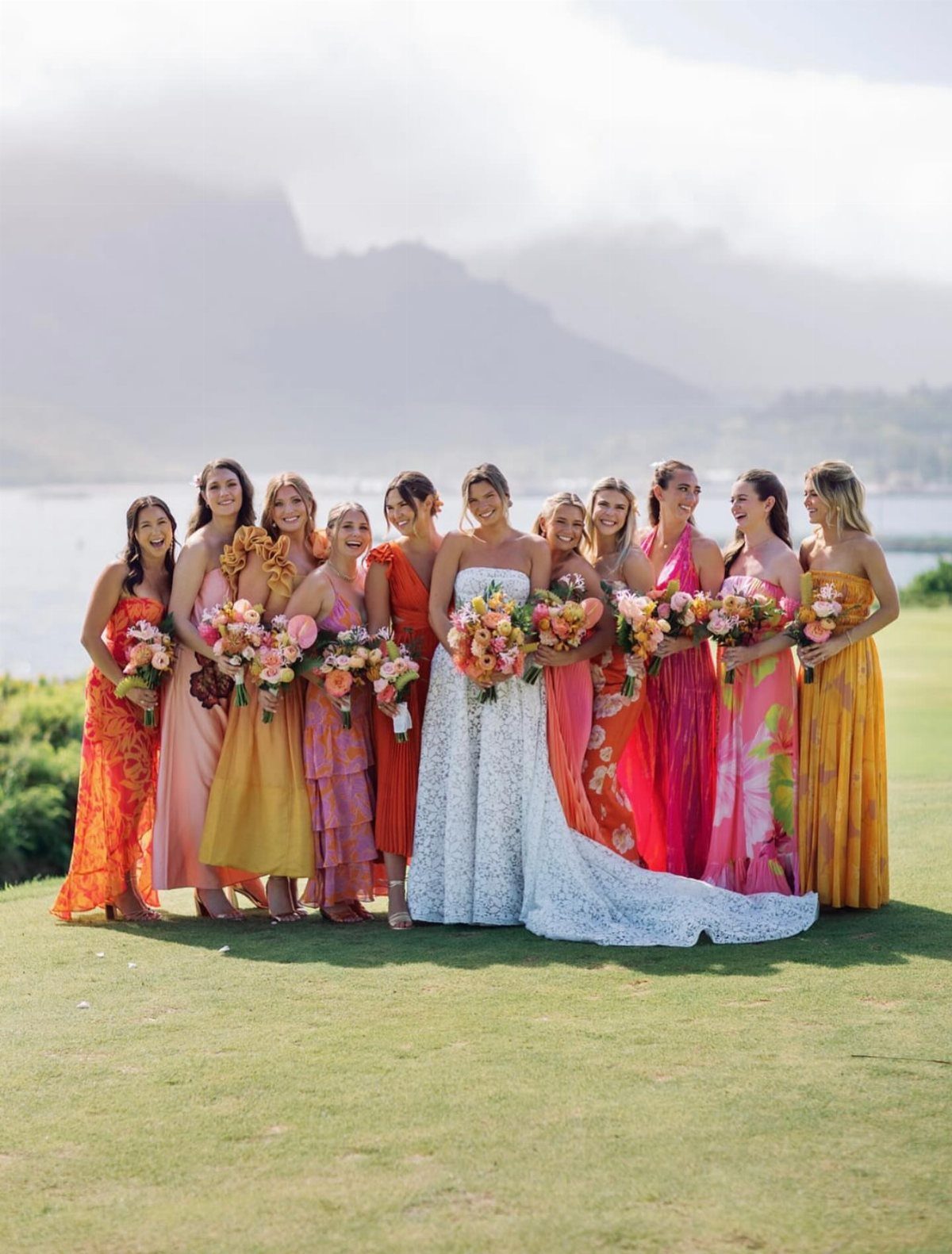 Bride and bridesmaids in colorful dresses holding tropical bouquets on a grassy lawn.