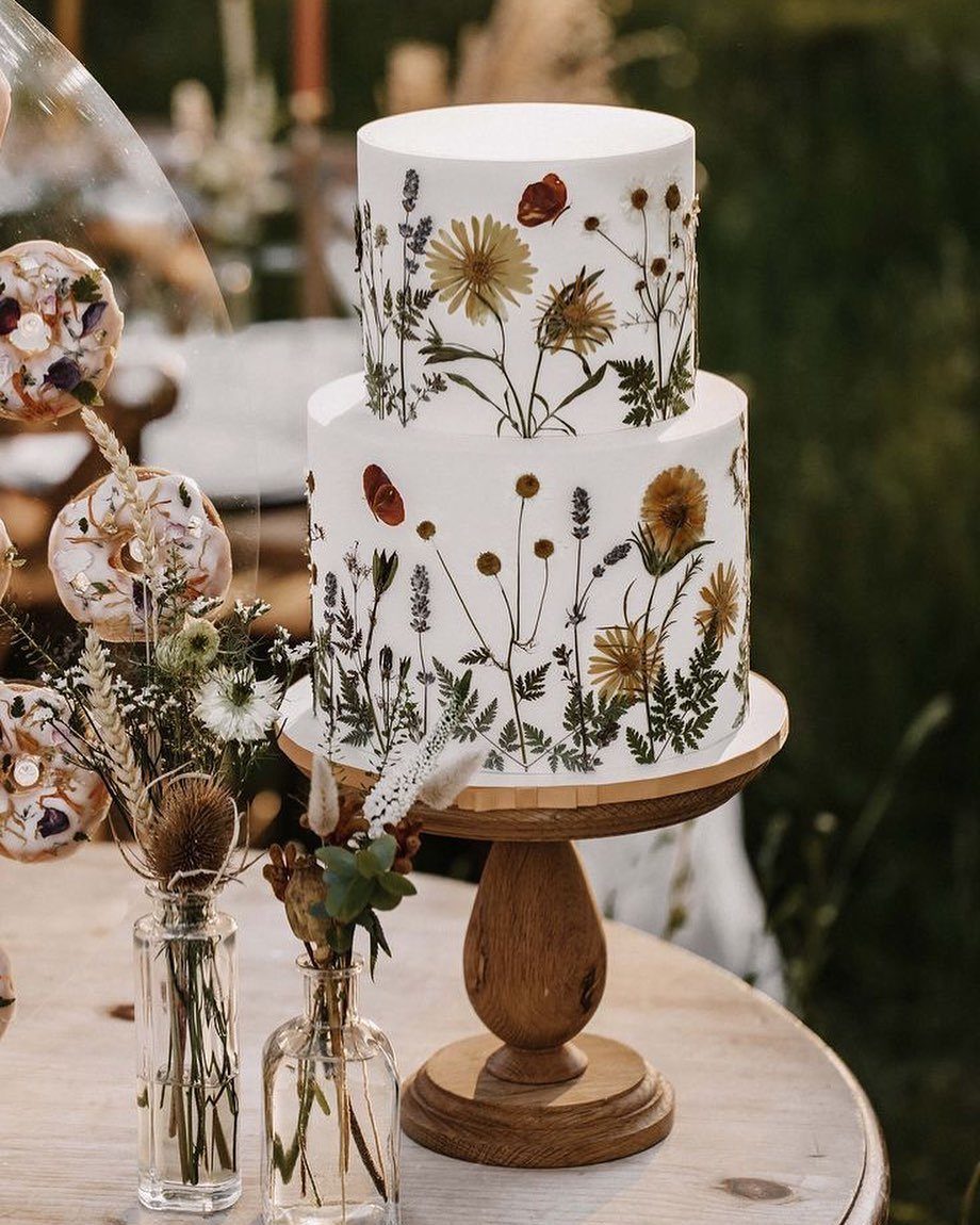 Two-tier white cake decorated with pressed dried flowers on a wooden stand.