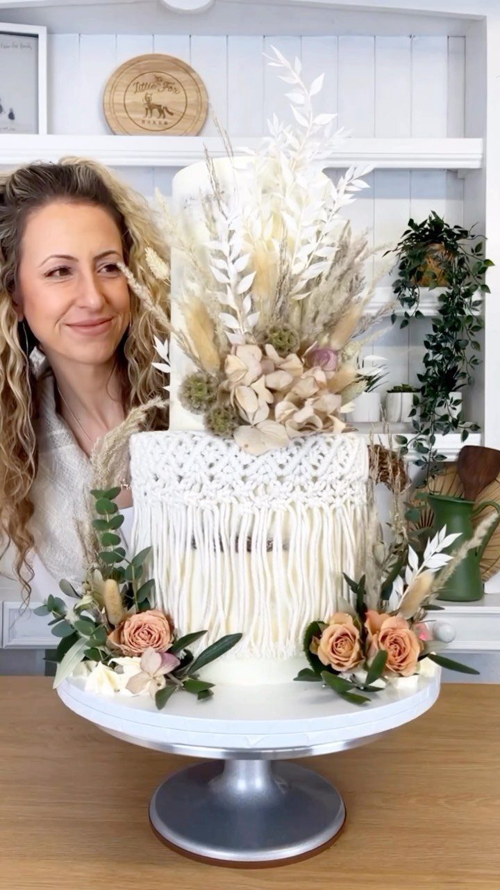 White tiered cake decorated with macramé lace, dried pampas grass, and peach roses.