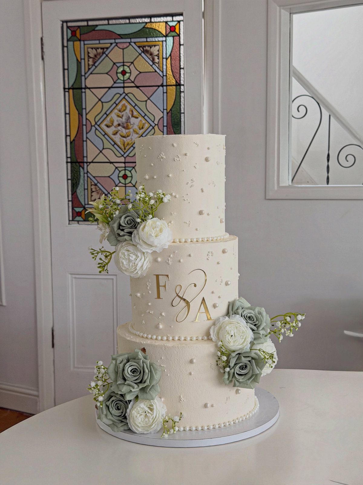 Three-tier white wedding cake with gold metallic initials and sage green roses.