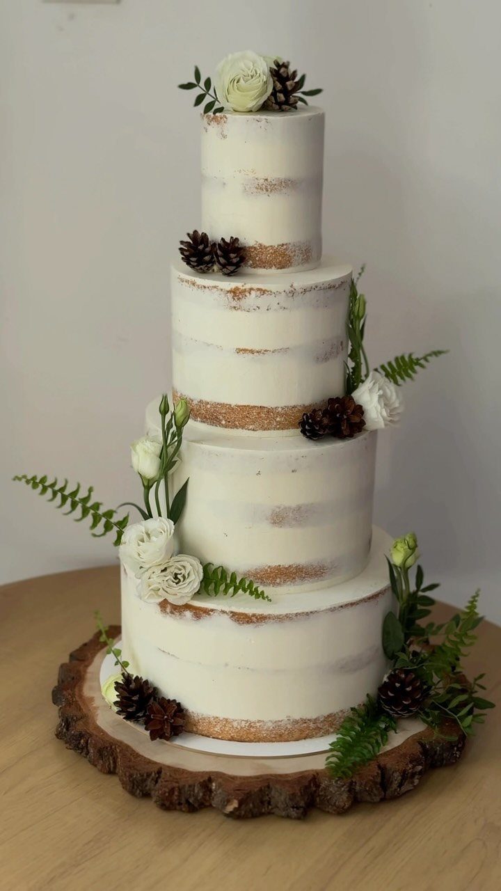 Four-tier semi-naked cake decorated with white roses and pinecones on a wood slice.