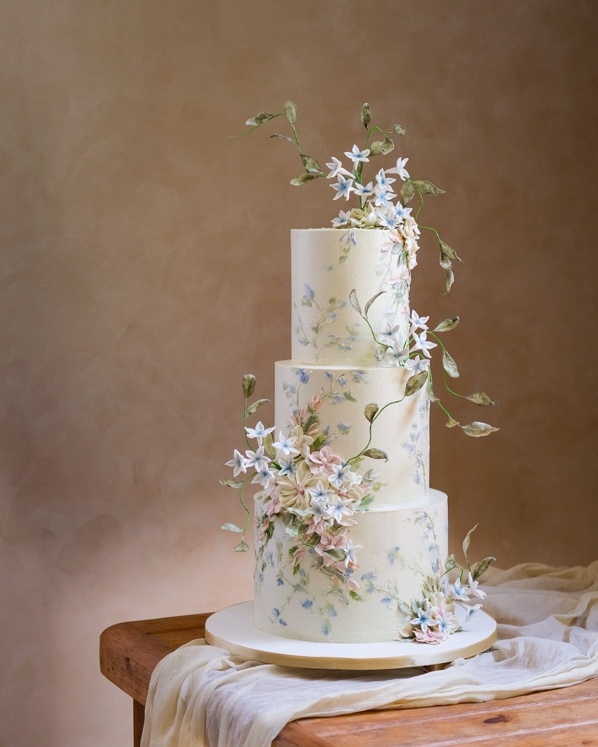 Three-tier white wedding cake with blue watercolor floral paintings and delicate 3D blossoms.