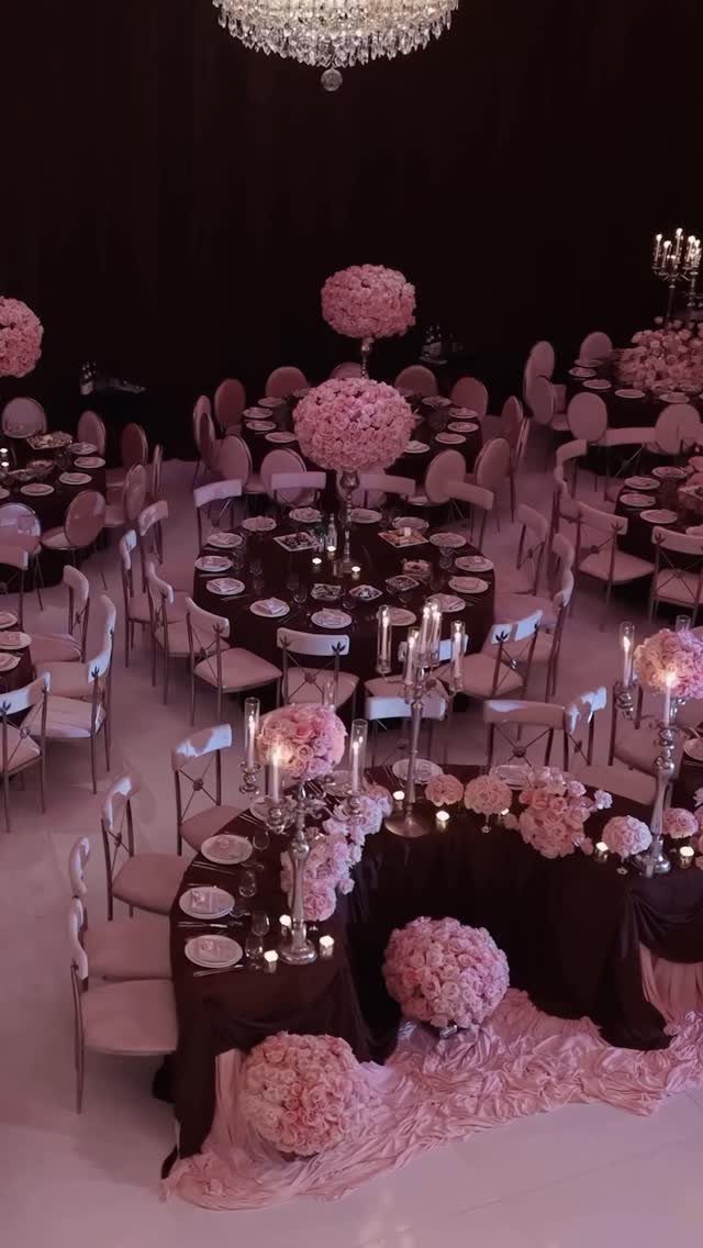 Wedding reception tables with burgundy linens and large pink floral centerpieces under a chandelier.