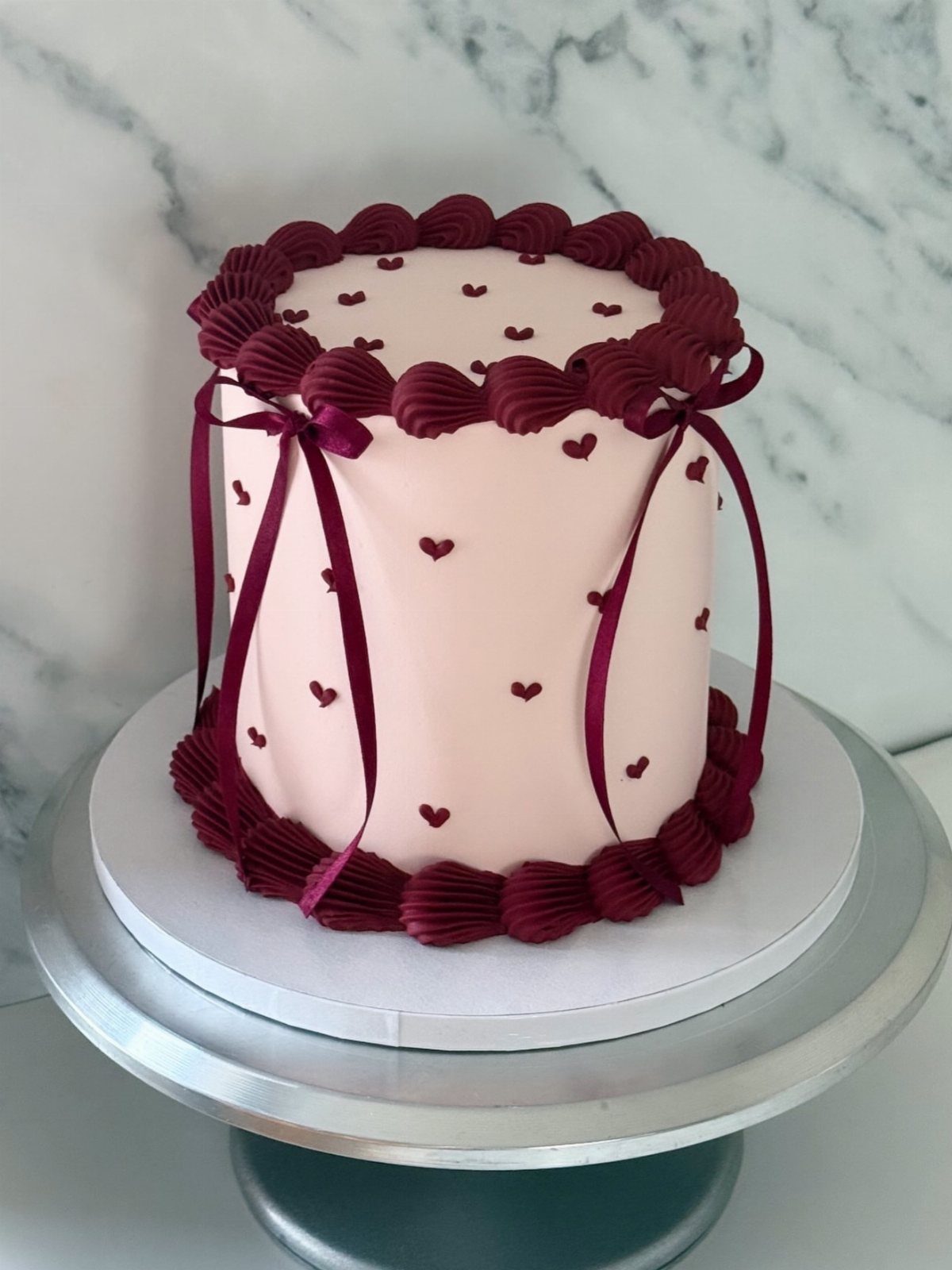 Pink cake with burgundy frosting swirls, ribbons, and hearts on a silver stand.