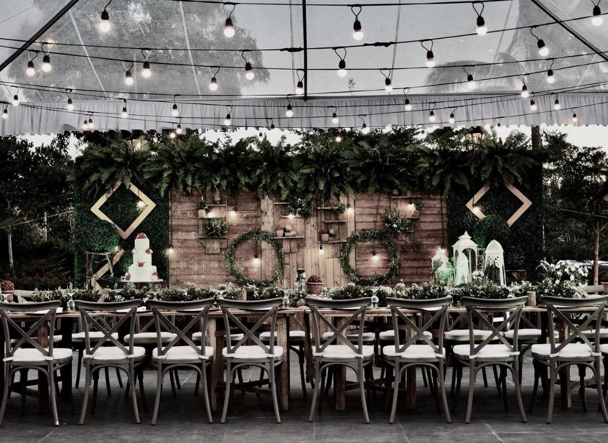 Wooden dining table and cross-back chairs under a clear tent with rustic greenery decor.