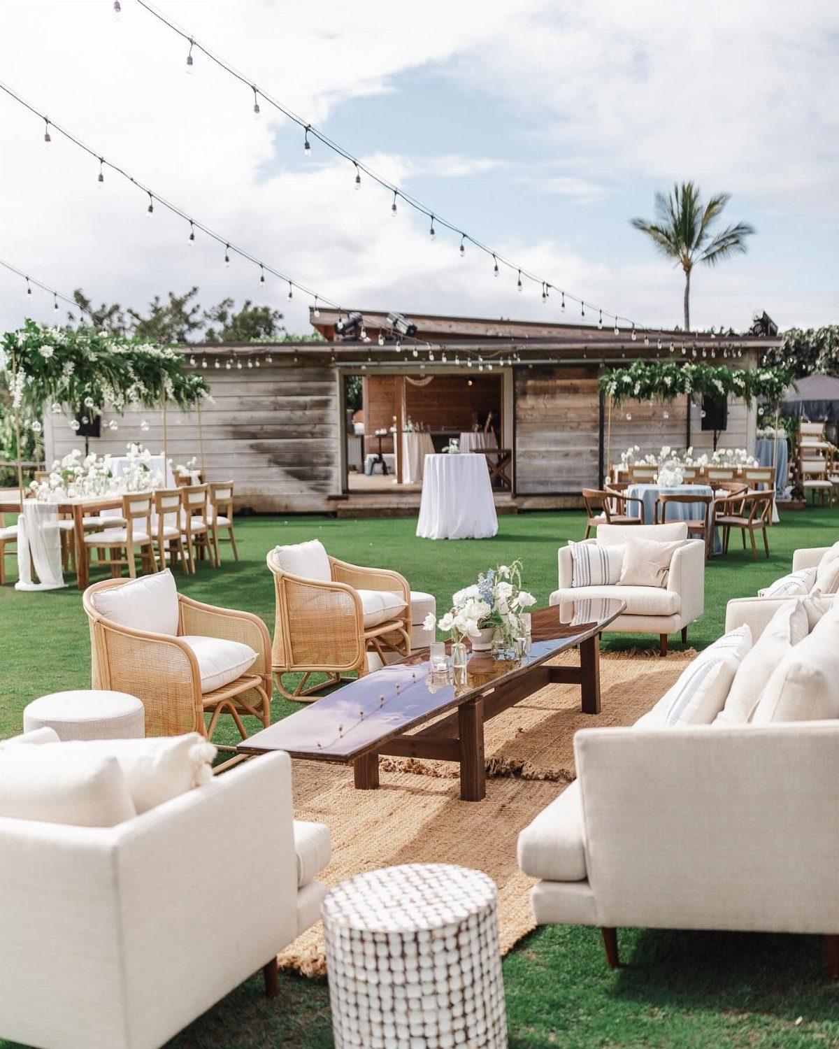 Outdoor wedding reception lounge with white sofas, wicker chairs, and rustic wooden building.
