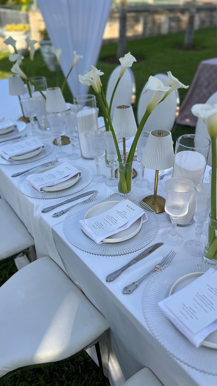 White wedding reception table setting with calla lilies and lamps on a green lawn