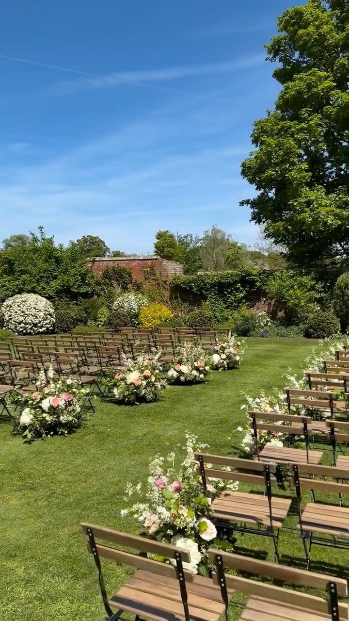 Wooden folding chairs and floral arrangements on a green lawn for an outdoor ceremony.