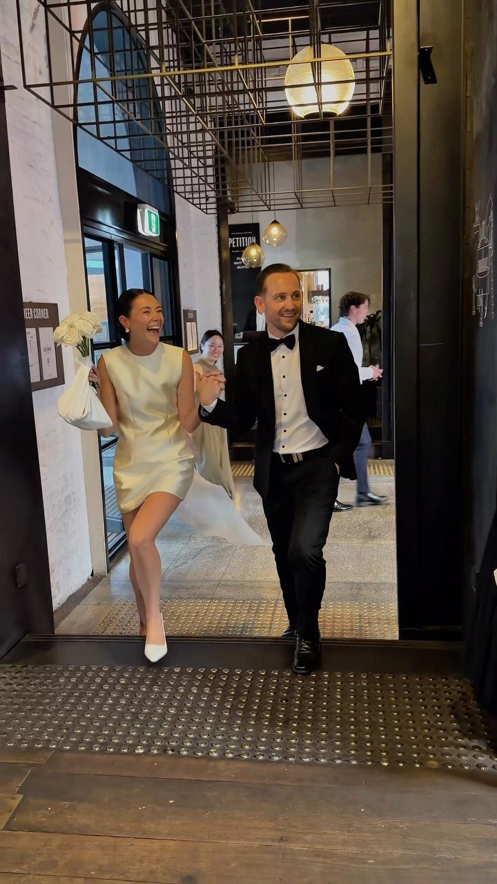 Bride in short white dress and groom in tuxedo walking through a doorway.