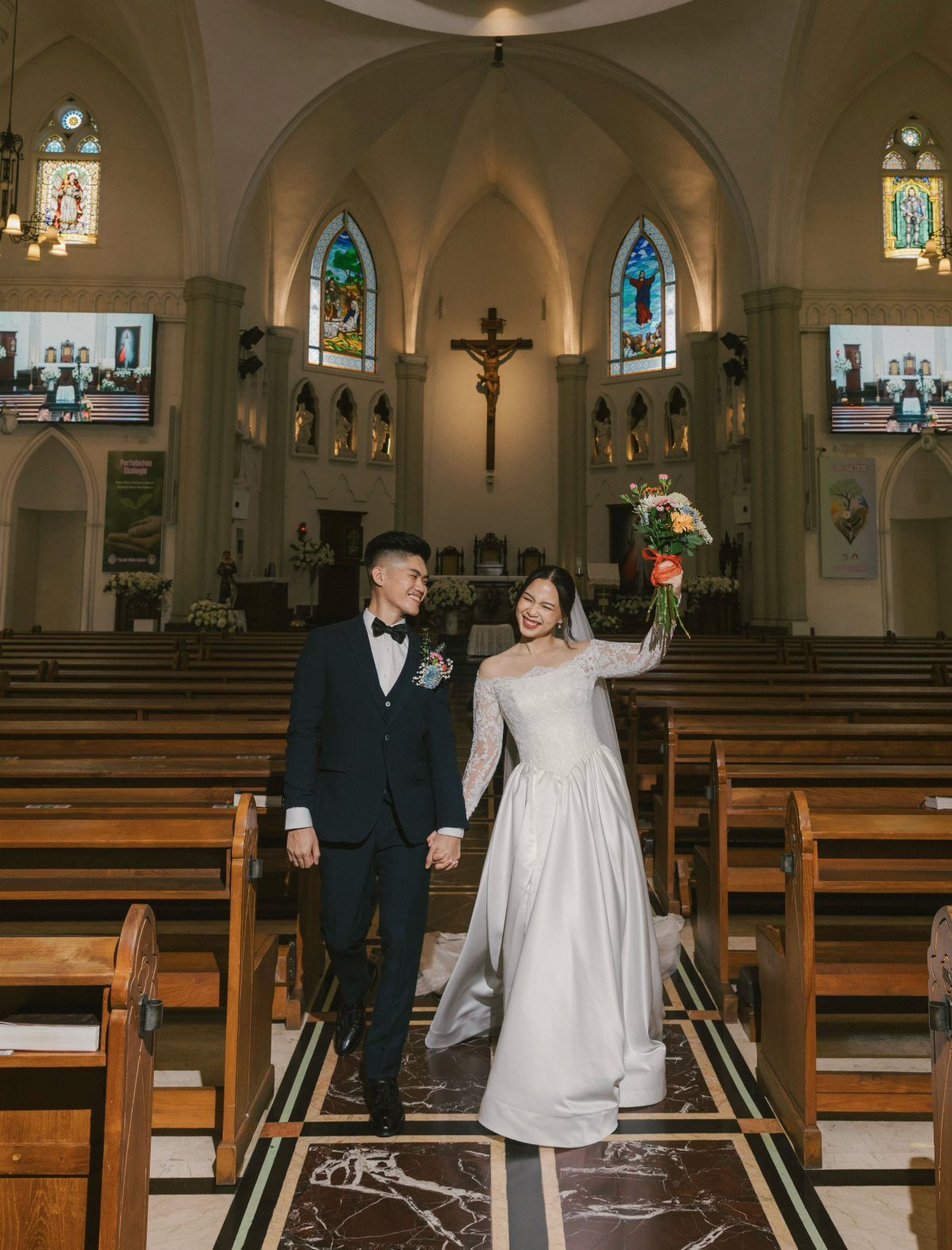 Bride in white gown and groom in navy tuxedo walking through a church aisle.