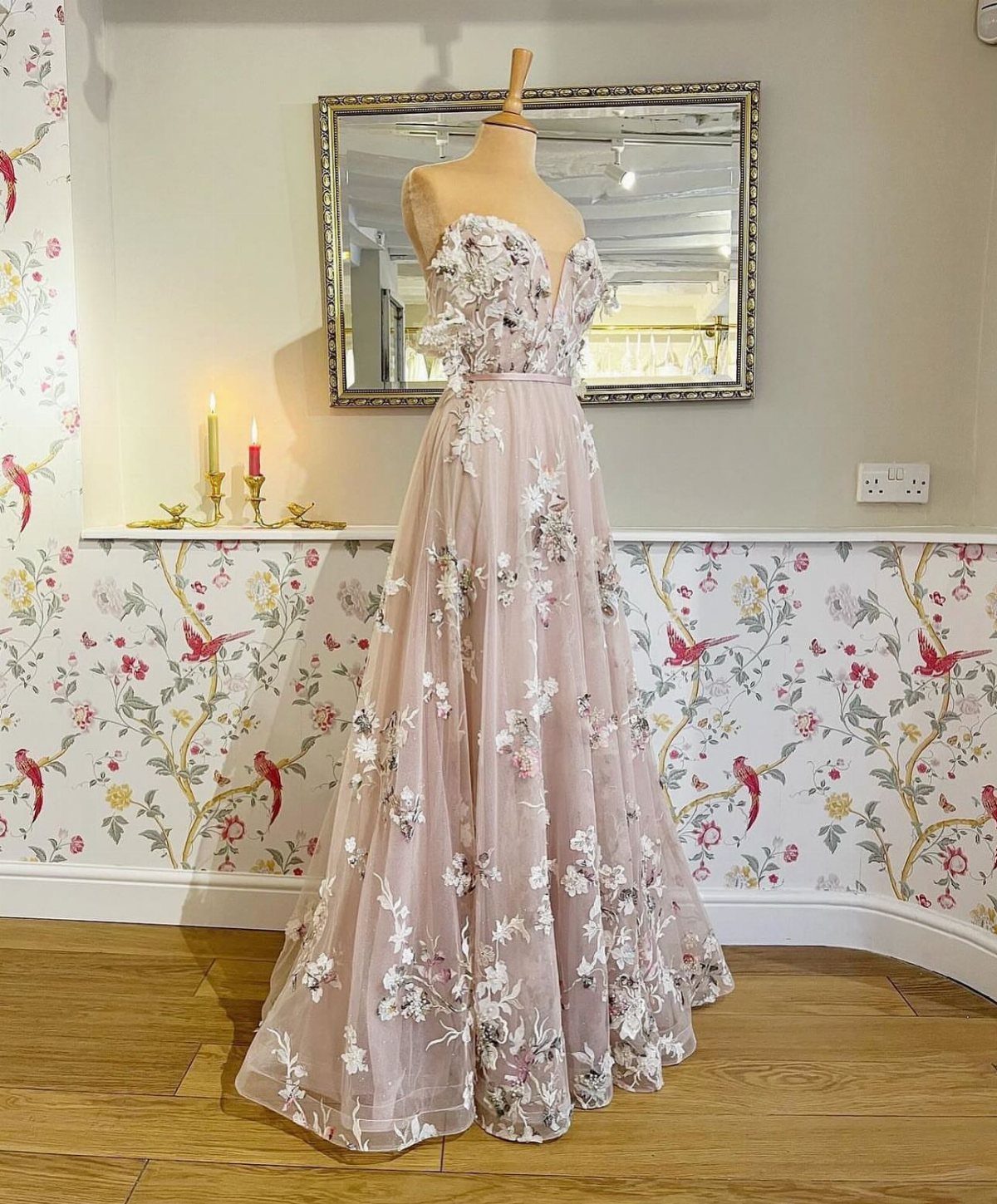 Blush-pink wedding dress with white floral embroidery on a mannequin in a room.
