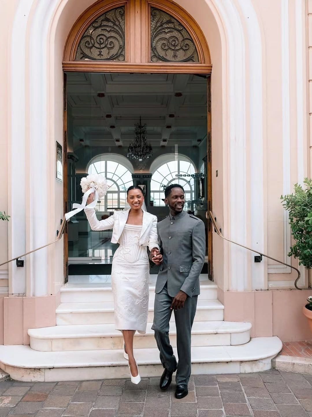 9 Court Wedding Outfit Ideas, From Jumpsuits to Mini Dresses 7 Couple in white dress and grey suit posing before an arched doorway.