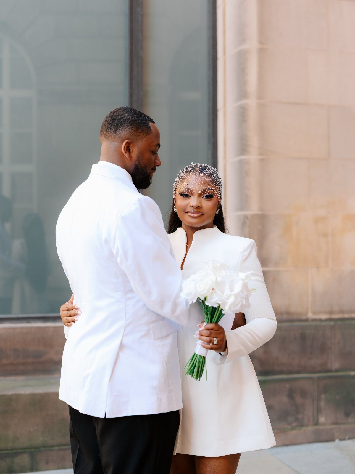 9 Court Wedding Outfit Ideas, From Jumpsuits to Mini Dresses 9 Couple in white wedding outfits, woman holding white flowers against a stone building.
