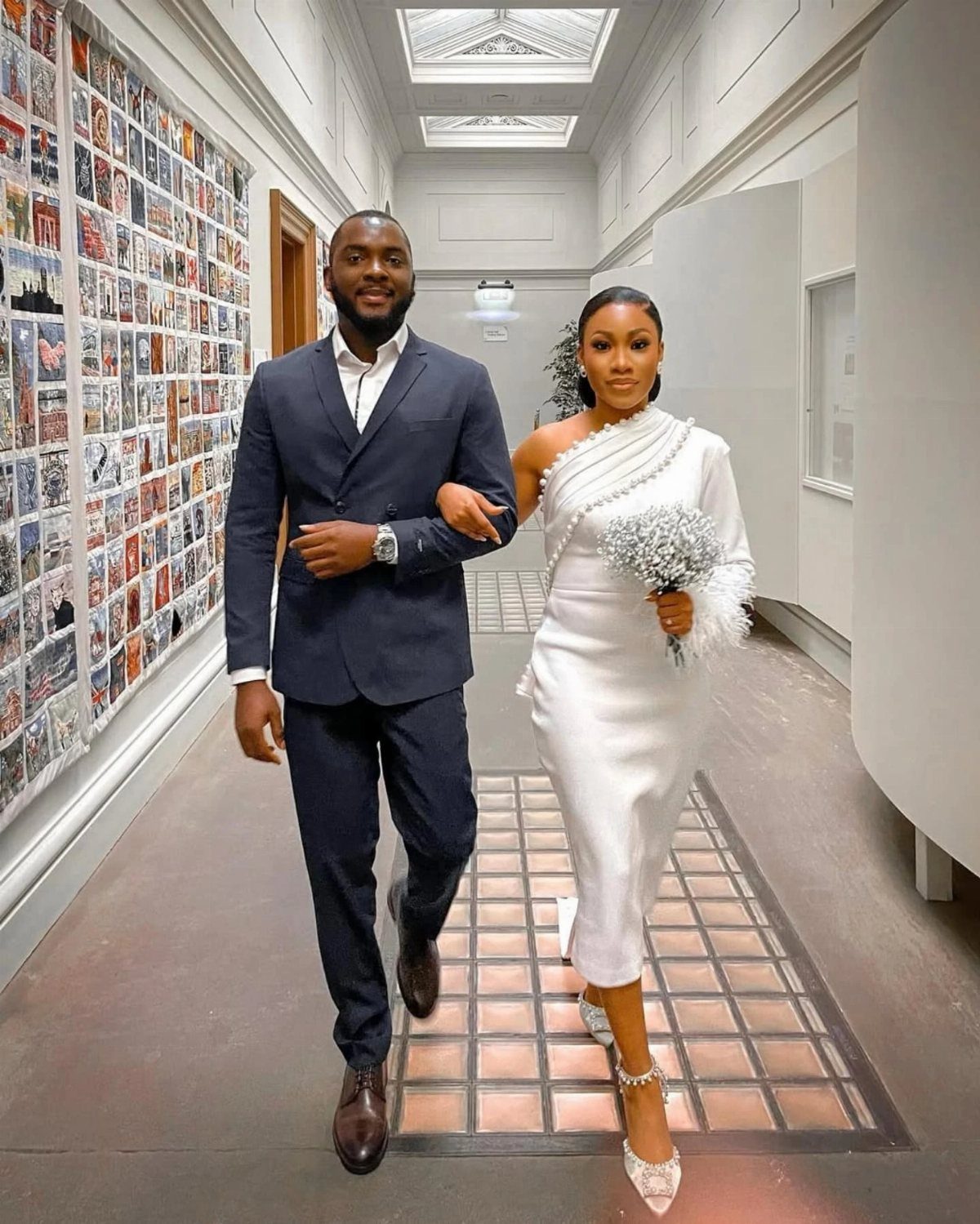 9 Court Wedding Outfit Ideas, From Jumpsuits to Mini Dresses 8 Couple in wedding attire walking through a gallery hallway, woman holding a white bouquet.