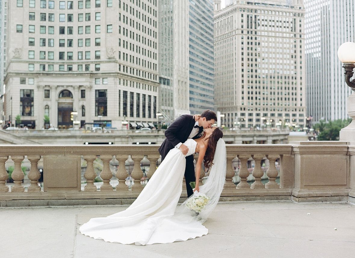 Groom dips bride in white wedding dress for a kiss on city bridge