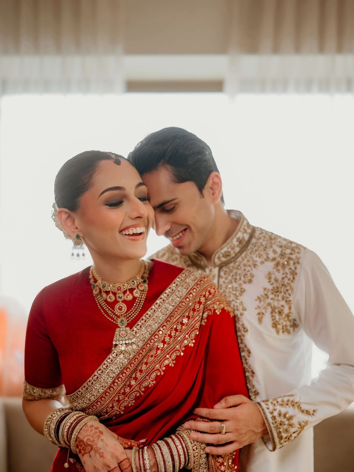 Bride in red saree and groom in white sherwani smiling closely together indoors.
