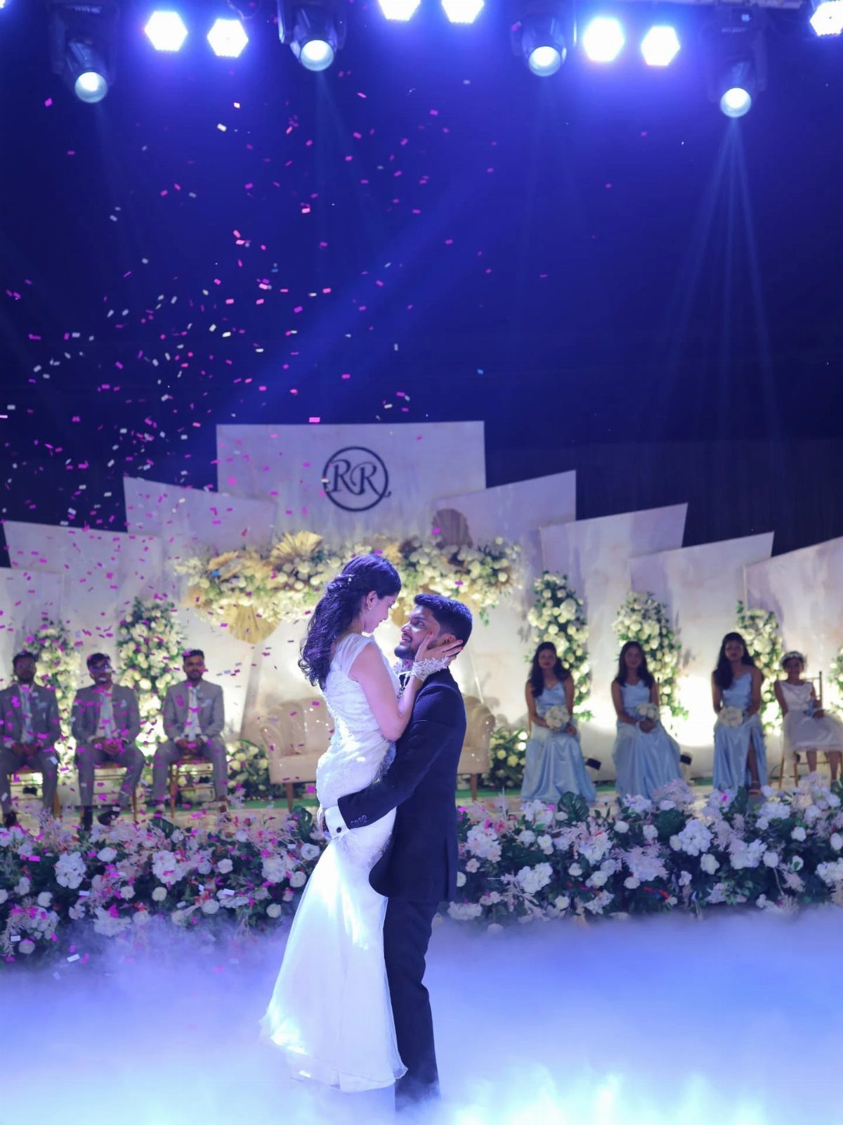 Bride and groom dancing together on a flower-decorated stage with blue lighting and confetti.
