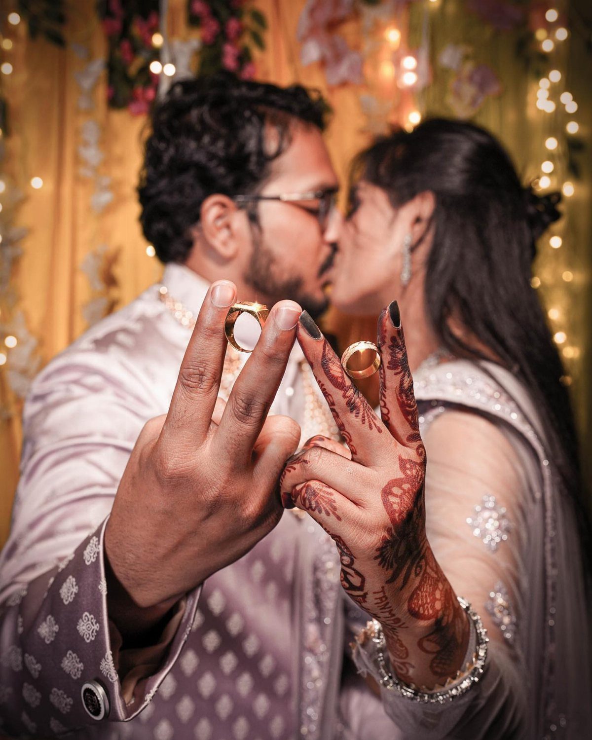 Couple kissing in background with wedding rings and henna hands in foreground focus.