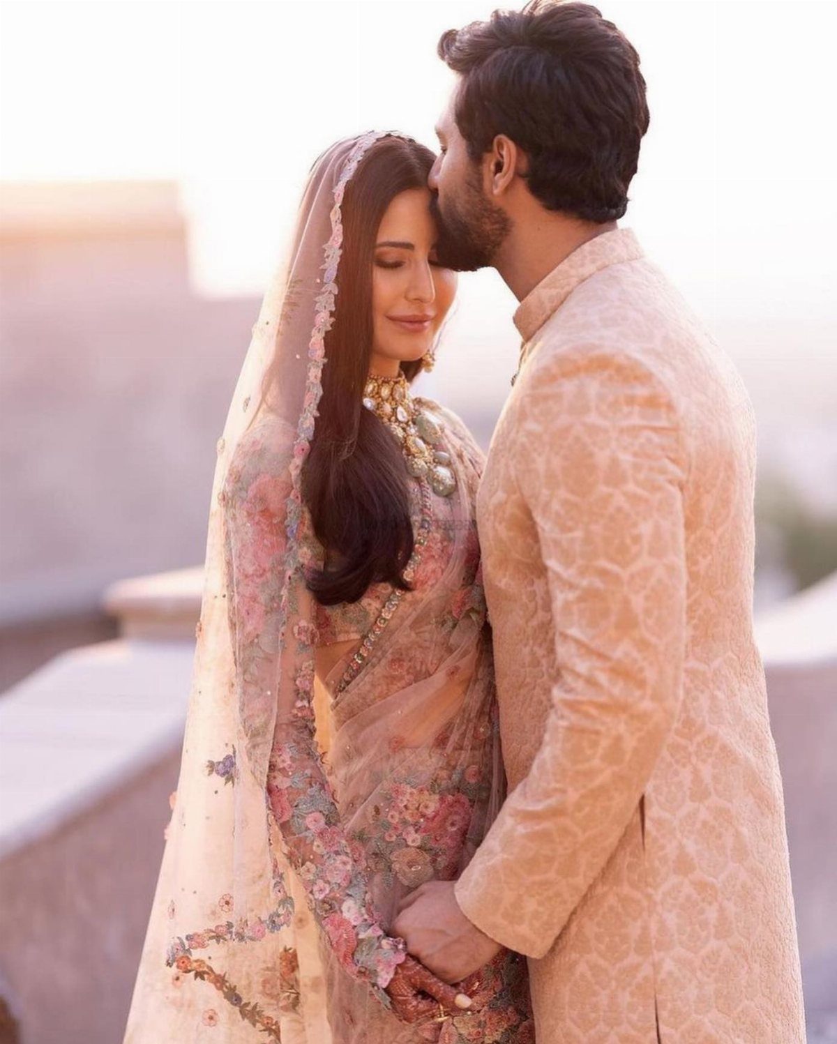 Groom in sherwani kissing bride on the forehead in traditional Indian wedding attire.