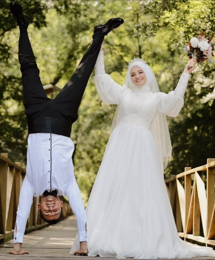 Bride in white dress smiling next to groom doing a handstand on bridge.