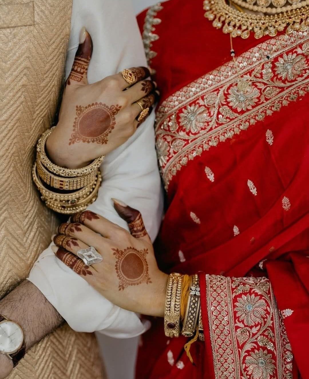 Henna-patterned hands of a bride in a red saree resting on a groom.