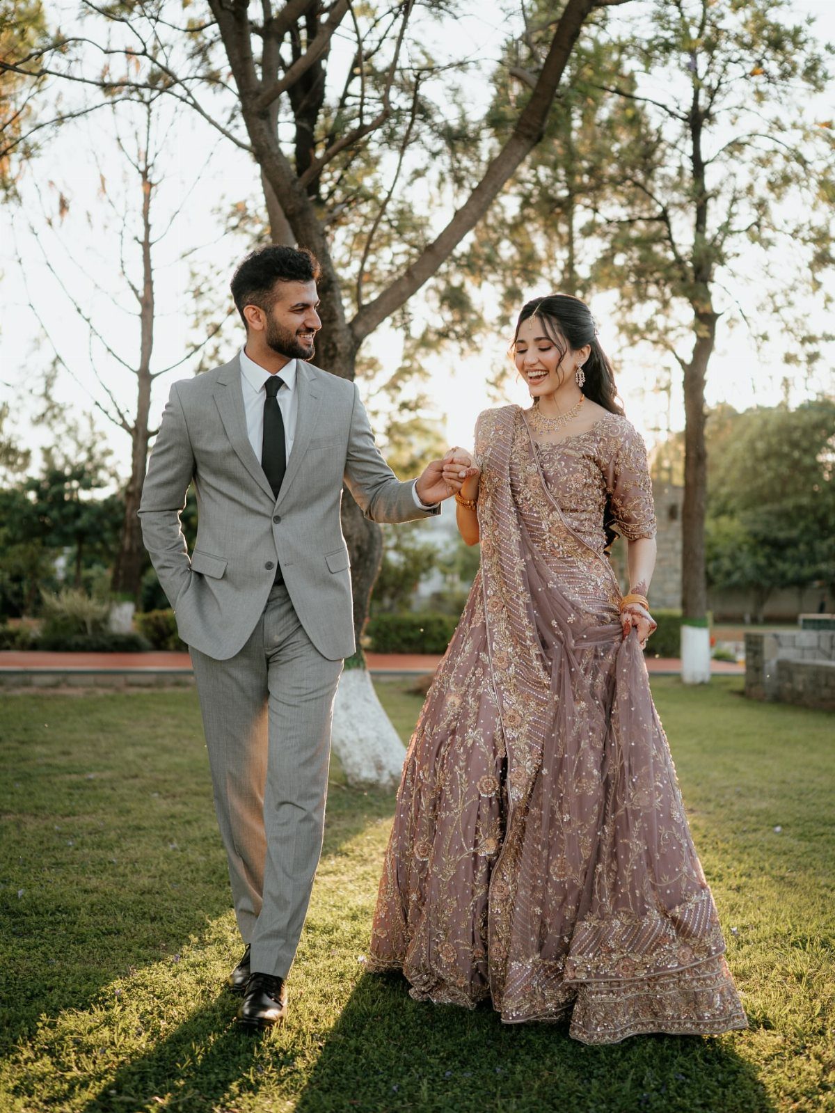 Couple in wedding attire holding hands and walking through a sunny park.