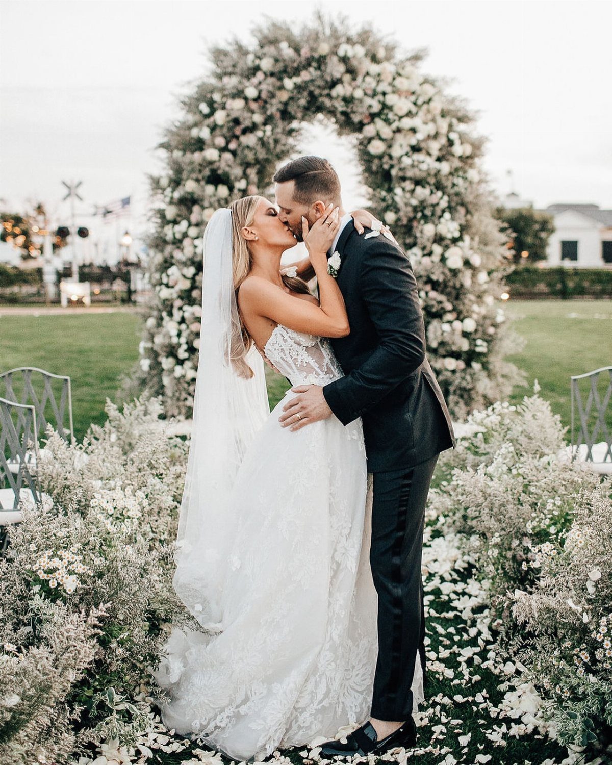 Bride and groom kissing in front of a large white floral wedding arch outdoors.