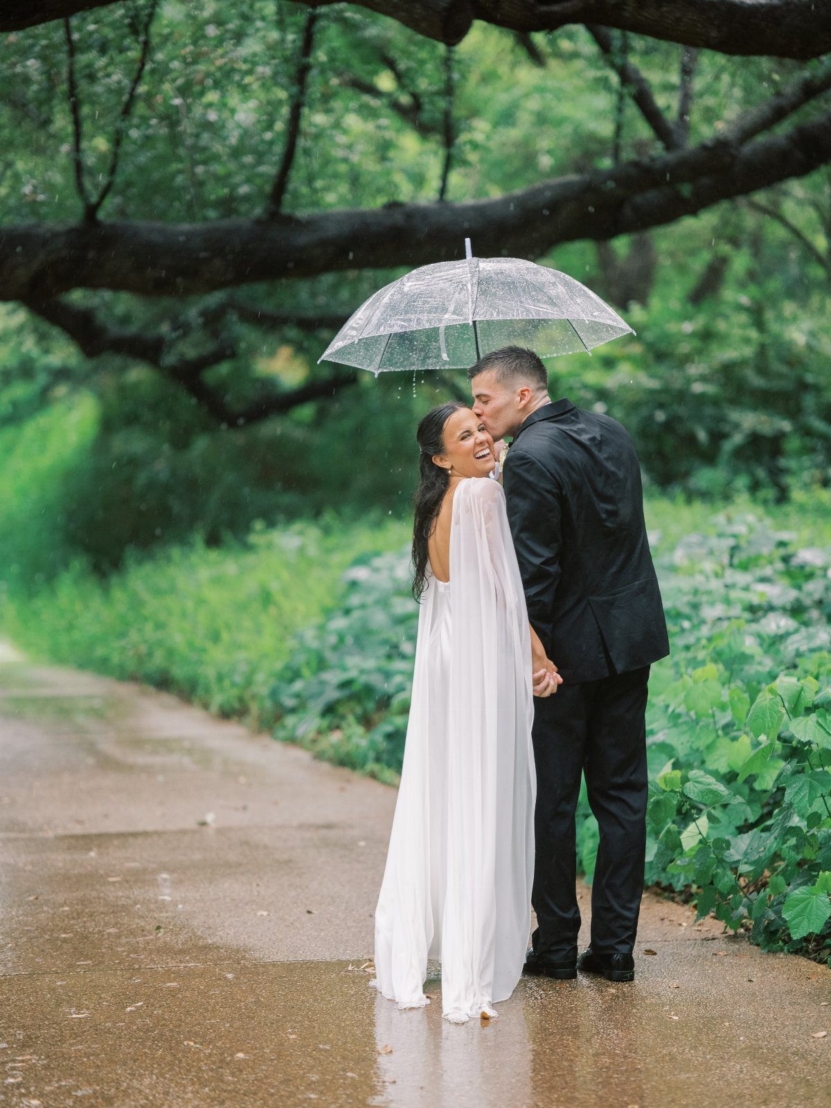 Bride and groom kissing under a clear umbrella on a rainy forest path.