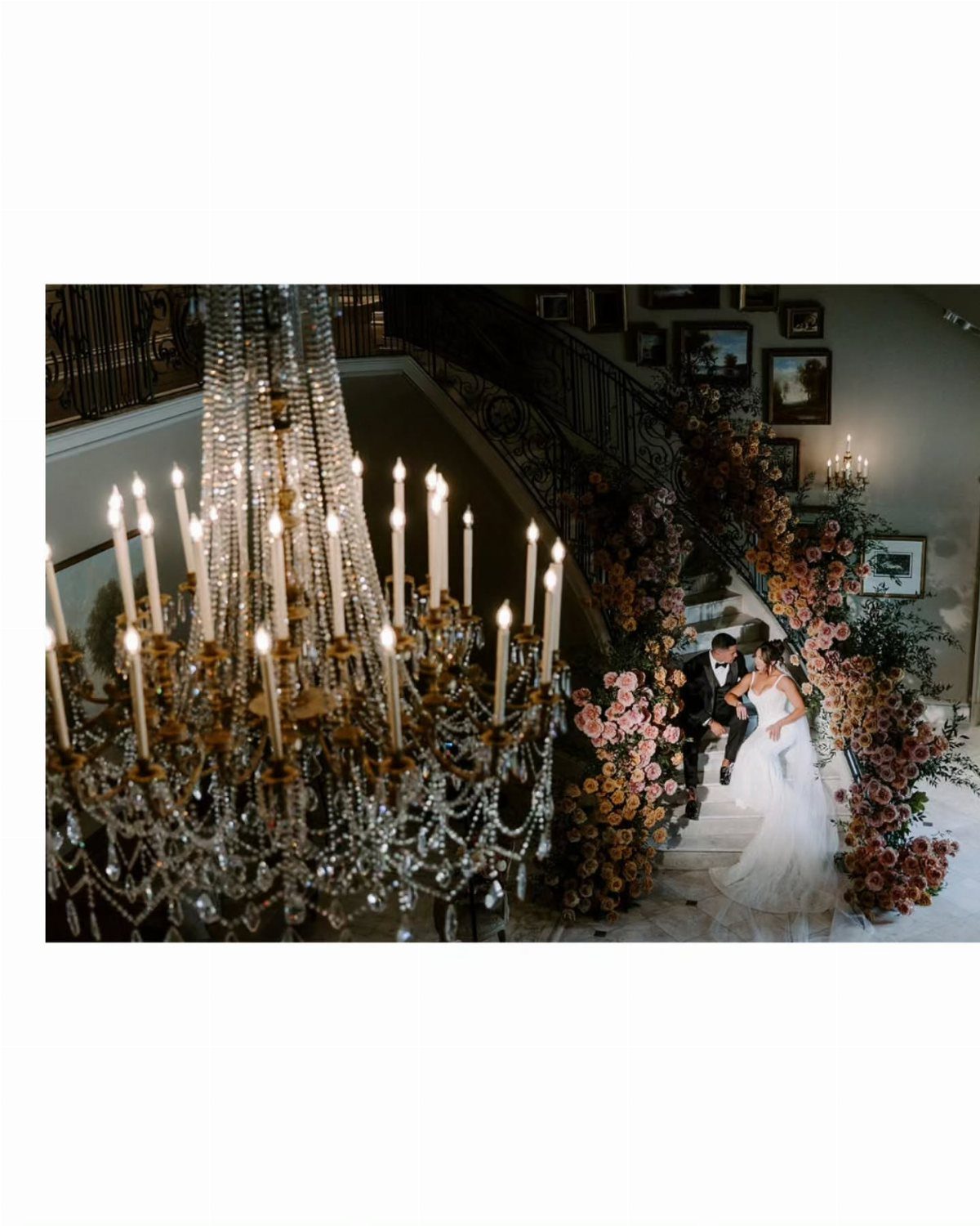 Bride and groom kissing on a floral staircase with a crystal chandelier foreground.