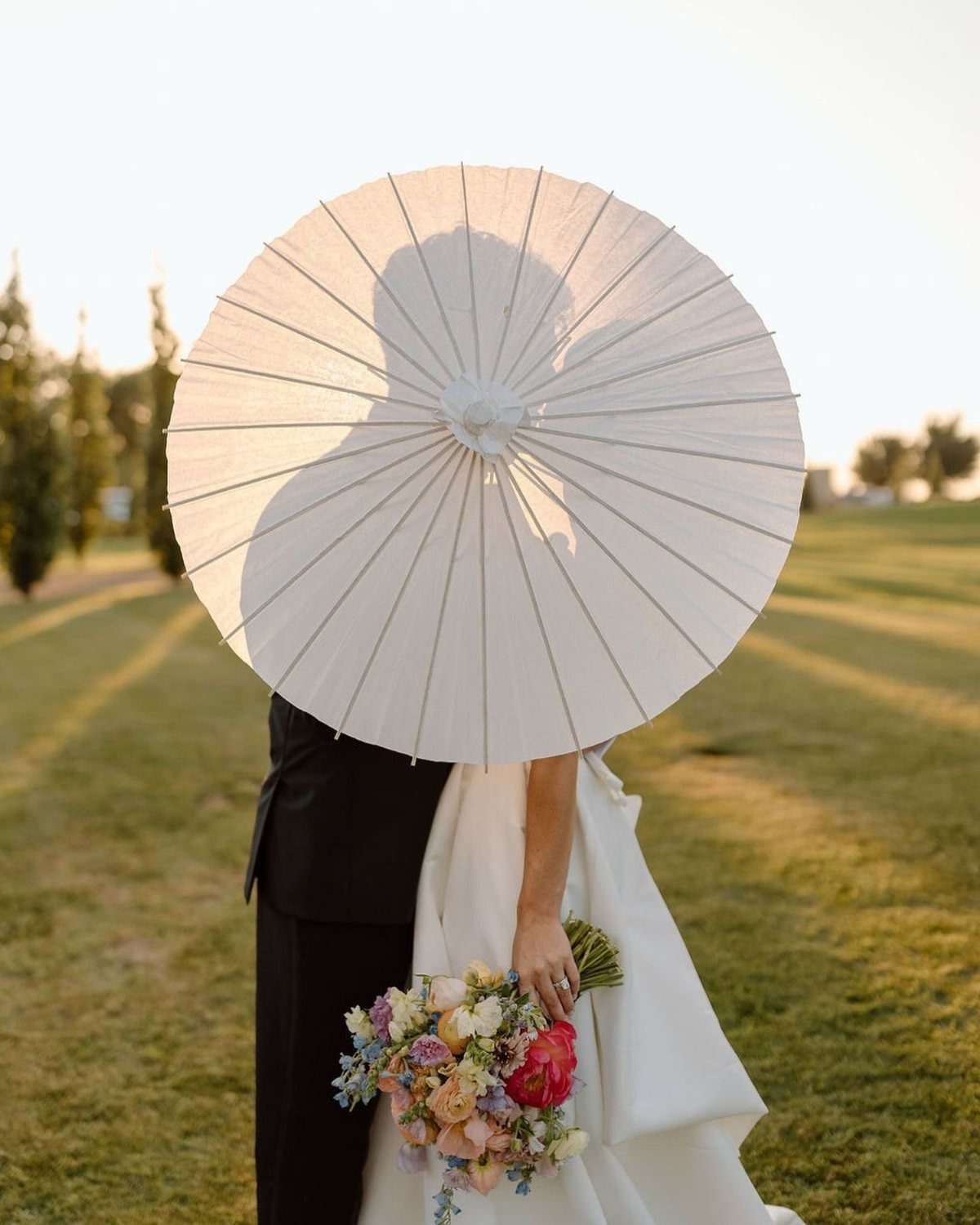 Bride and groom posing behind a white paper parasol in a sunny field