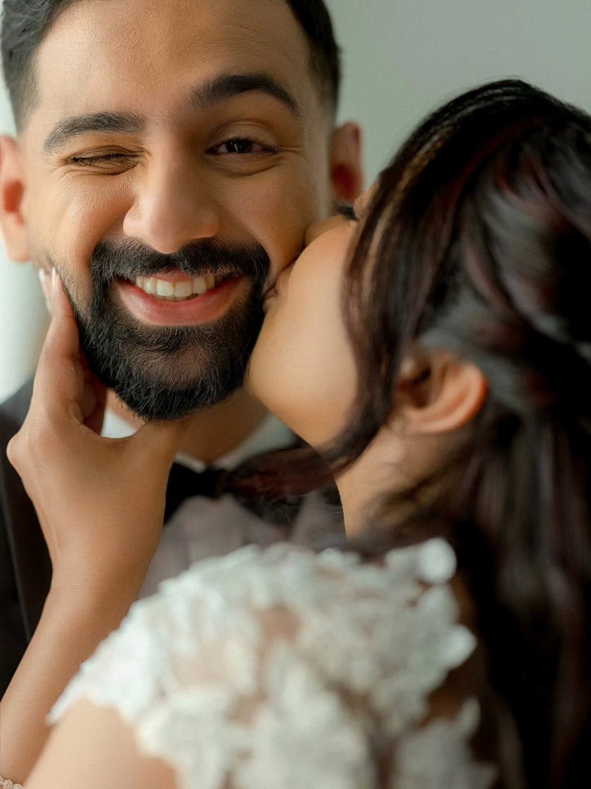 Groom winking and smiling while a bride in lace dress kisses his cheek.