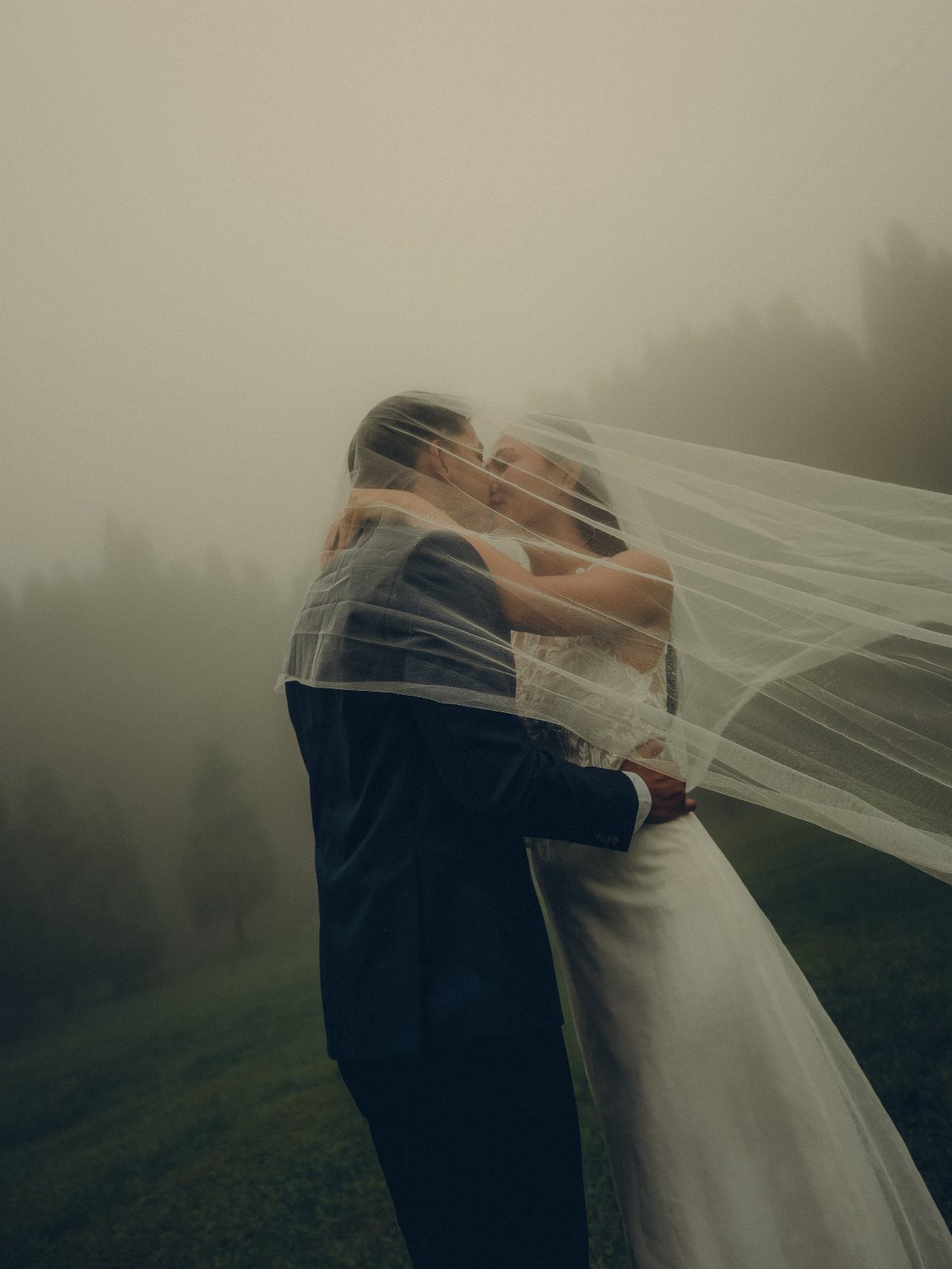 Bride and groom kissing while wrapped in a sheer white wedding veil in fog.