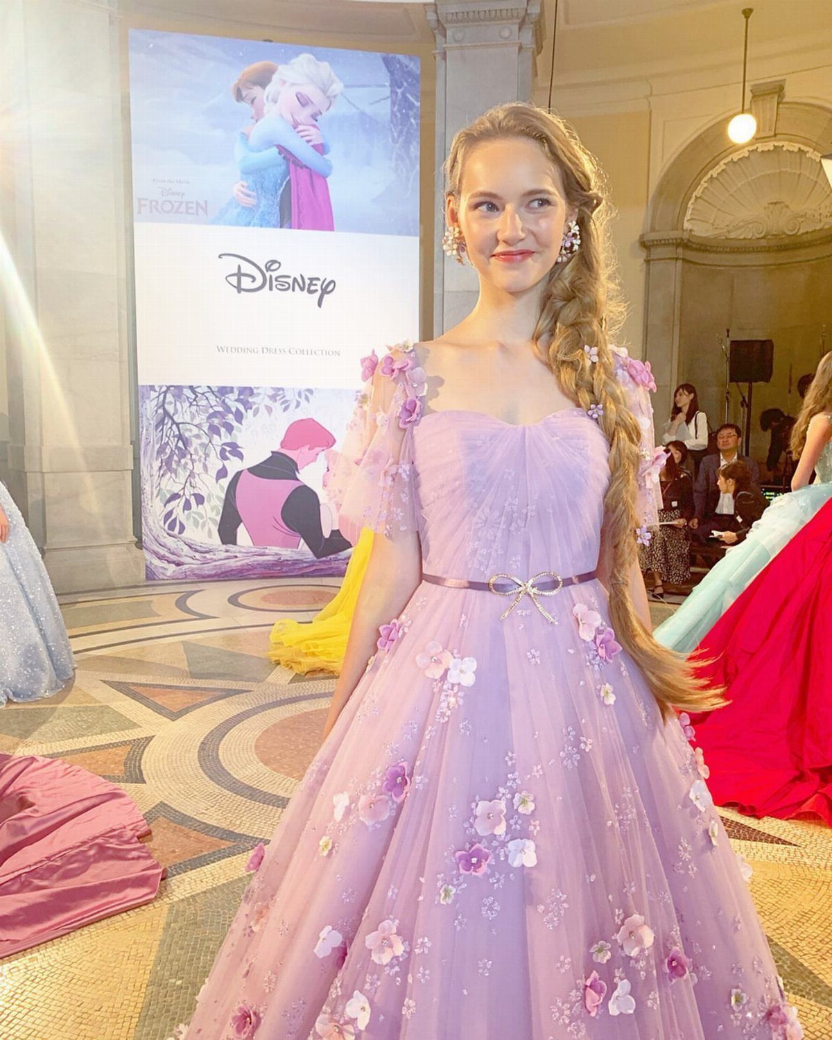 Woman in a purple floral gown with a long blonde braid standing indoors.