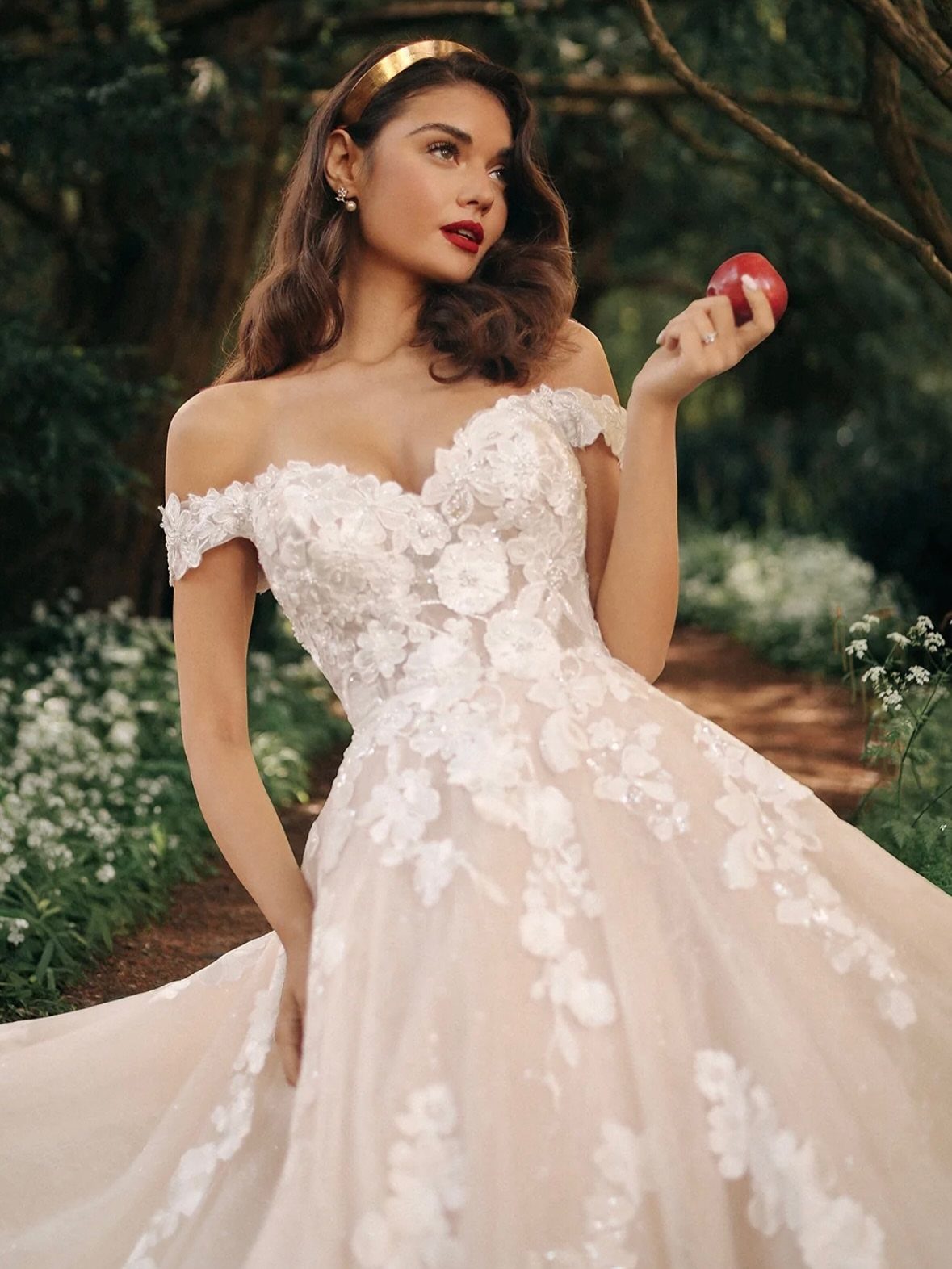 Woman in white floral wedding gown holding a red apple in a forest.