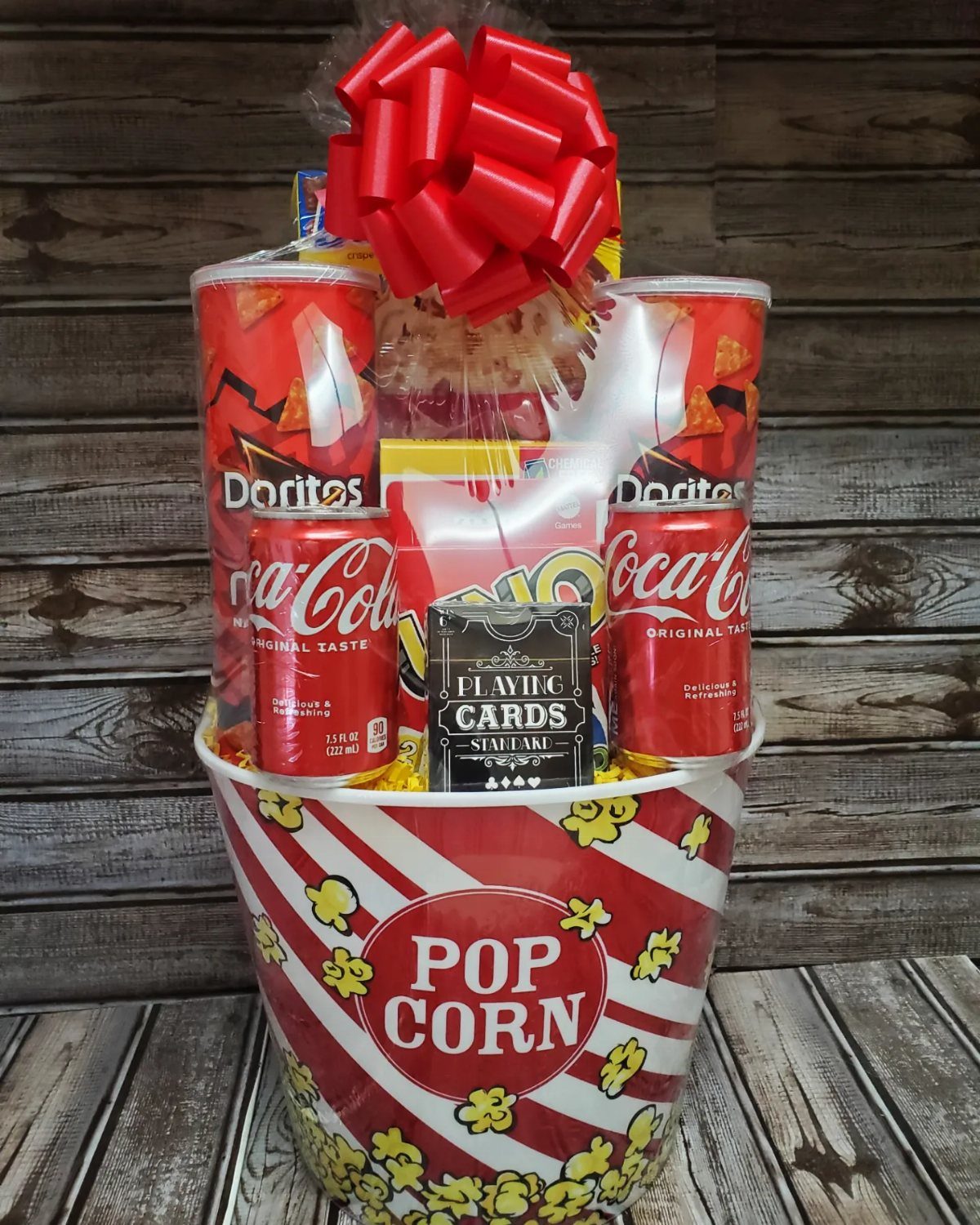 Popcorn bucket gift basket with soda, chips, and cards wrapped in cellophane with red bow.