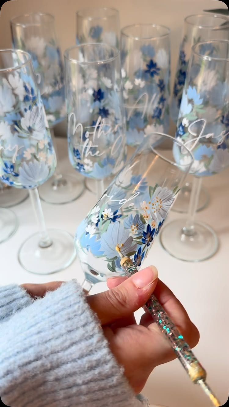 Hand painting blue floral designs on a champagne flute among other decorated glasses.