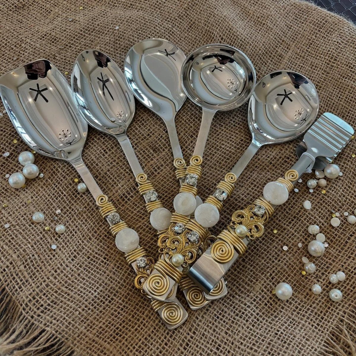 Silver serving utensils with gold wire wrapping and pearls on burlap fabric.