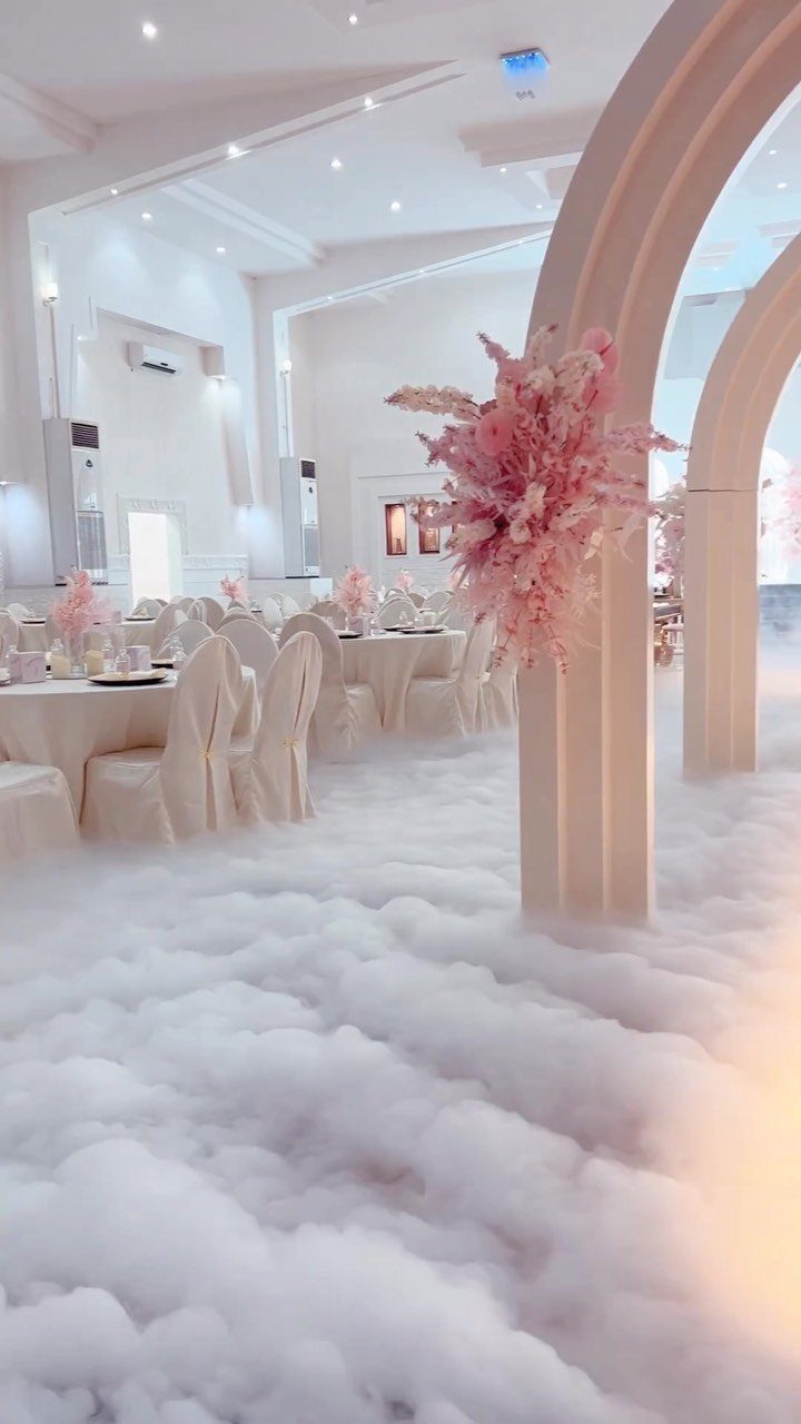 Wedding venue with white fog on the floor, white tables, and pink floral arches.