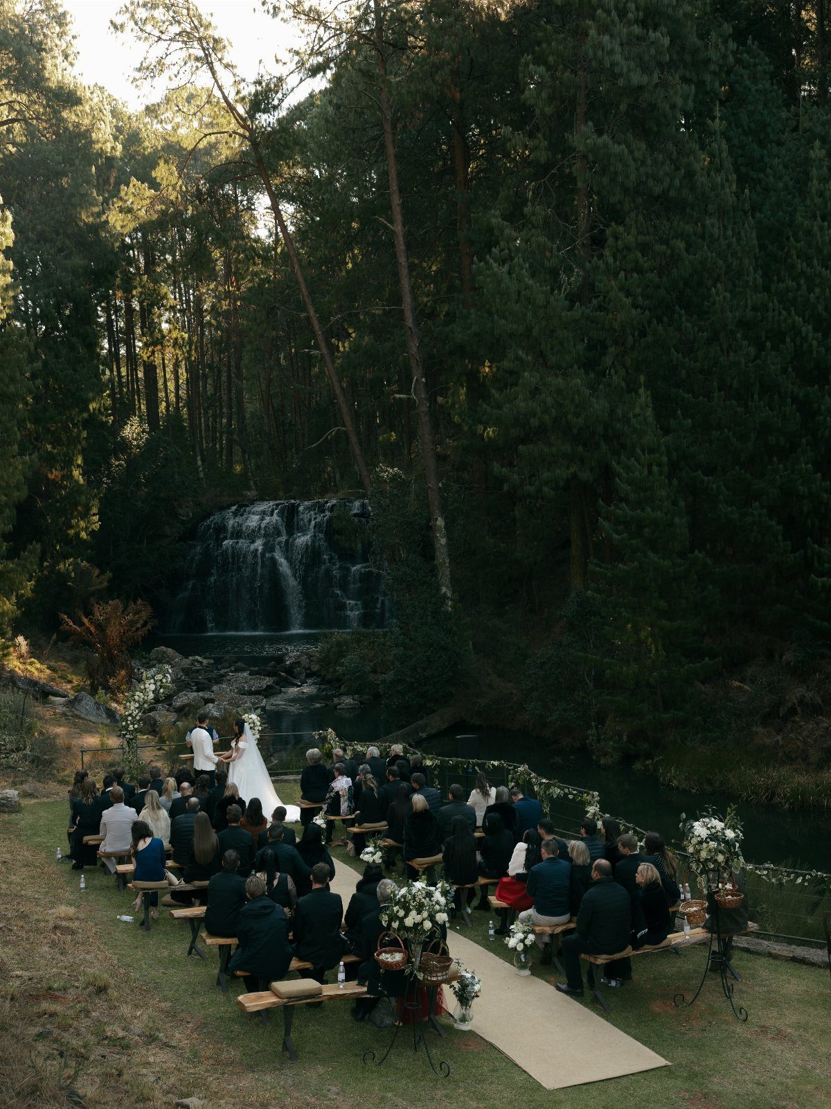 Wedding ceremony in a forest clearing with a waterfall and guests on benches.