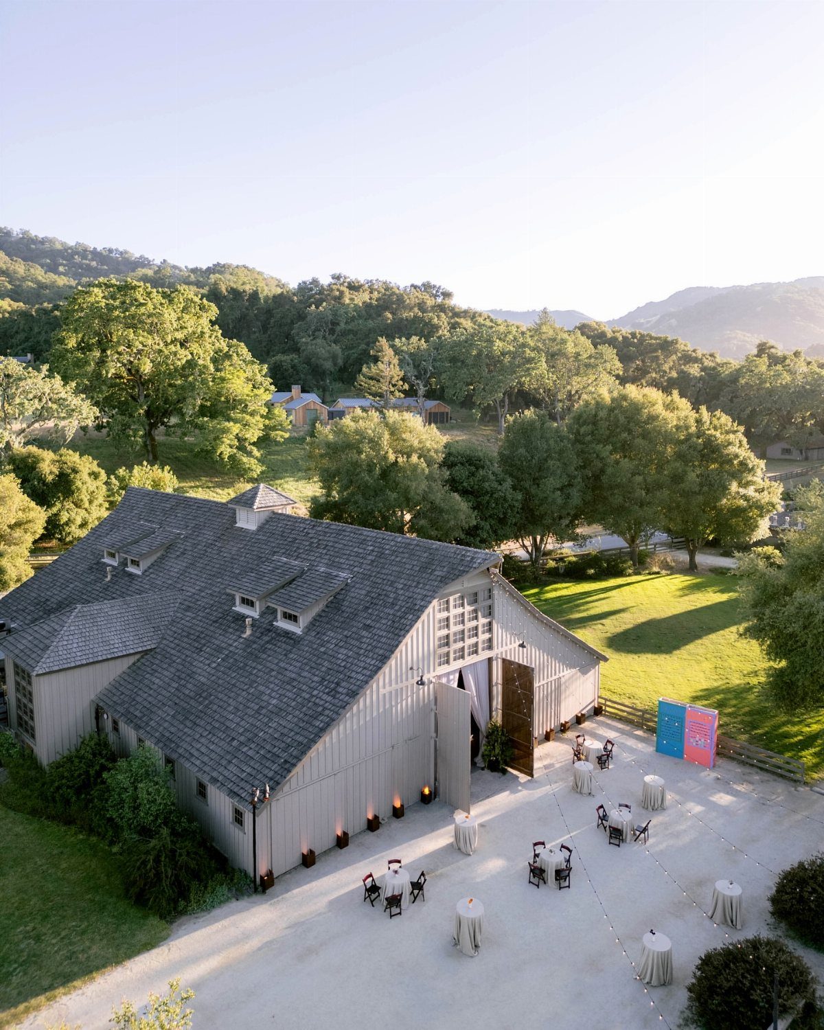 Grey barn wedding venue with outdoor cocktail tables set against rolling green hills.