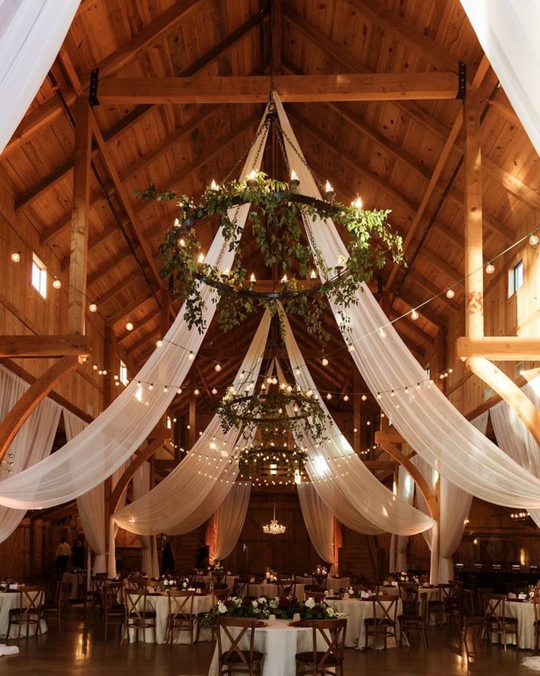 Dream Wedding Venue Ideas: 11 Places to Say I Do 4 Rustic wooden barn interior with white fabric draping, greenery chandeliers, and wedding reception tables.