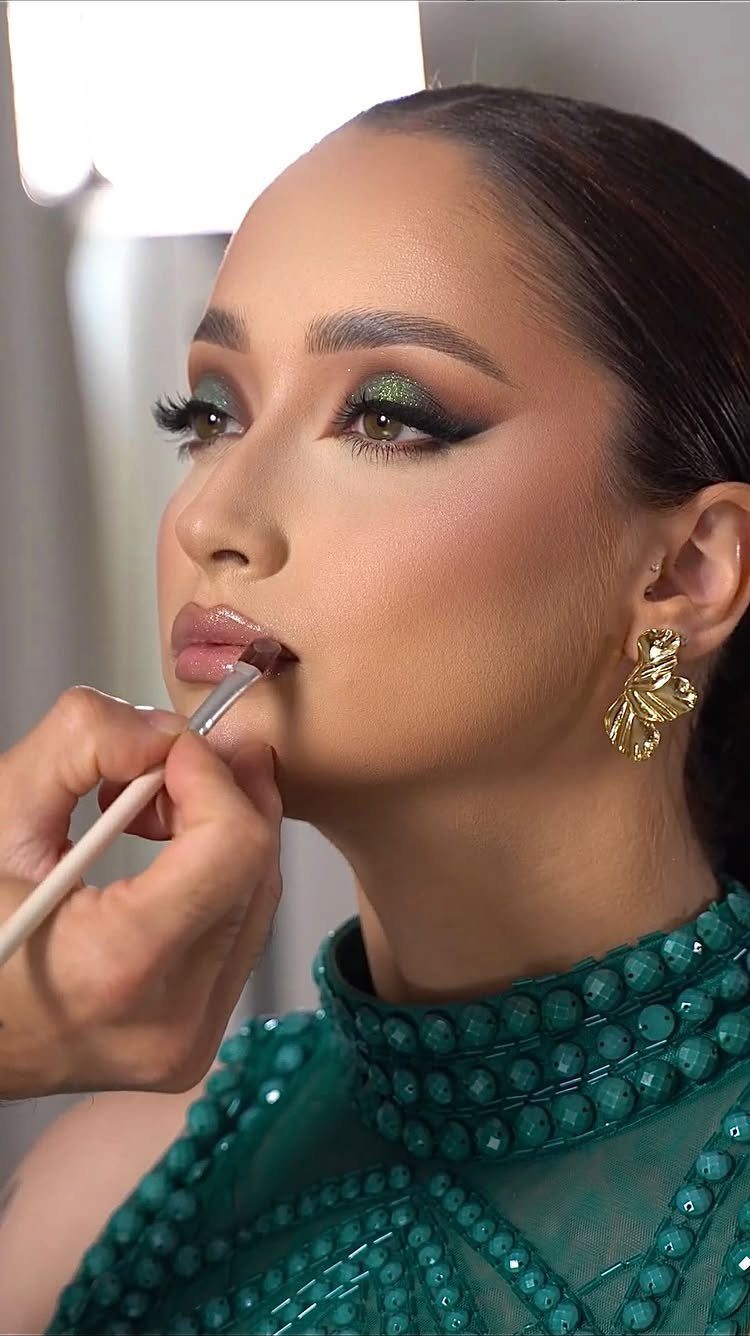 Woman with green shimmering eye makeup and a green beaded dress receiving lip application.