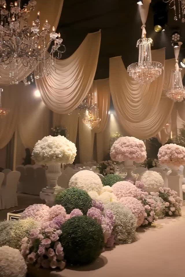 Crystal chandeliers and cream fabric drapery above lush white and pink floral arrangements.