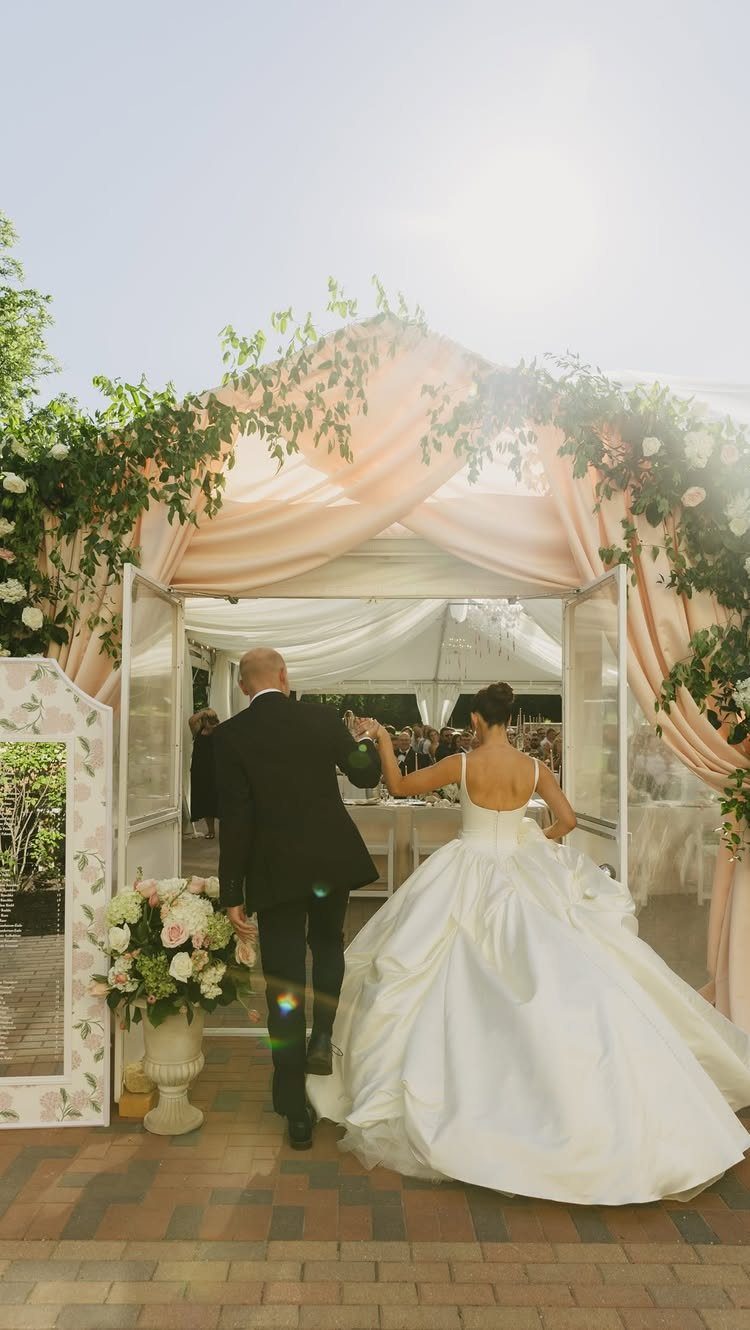 Bride and groom entering a wedding tent draped in peach fabric and greenery.